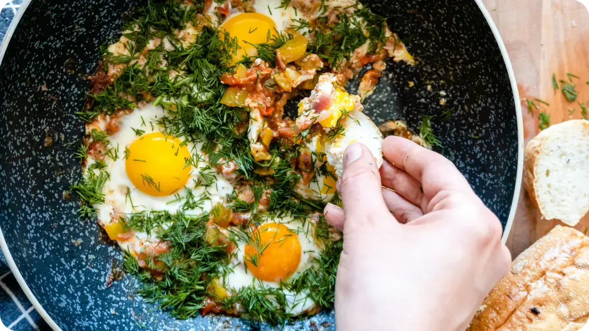 Three fried eggs covered in bacon and dill, with a hand dipping bread into a yolk.