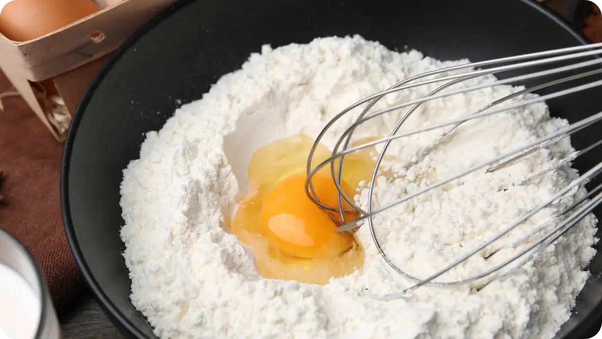A mixing bowl filled with flour and an egg, with a whisk resting to the side.