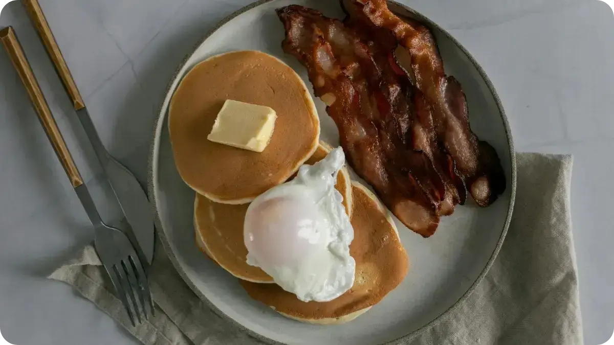 A plate of breakfast food with a stack of pancakes topped with butter and a poached egg, served alongside crispy bacon, with a fork and knife placed on a light tablecloth. A plate of breakfast food with a stack of pancakes topped with butter and a poached egg, served alongside crispy bacon, with a fork and knife placed on a light tablecloth.