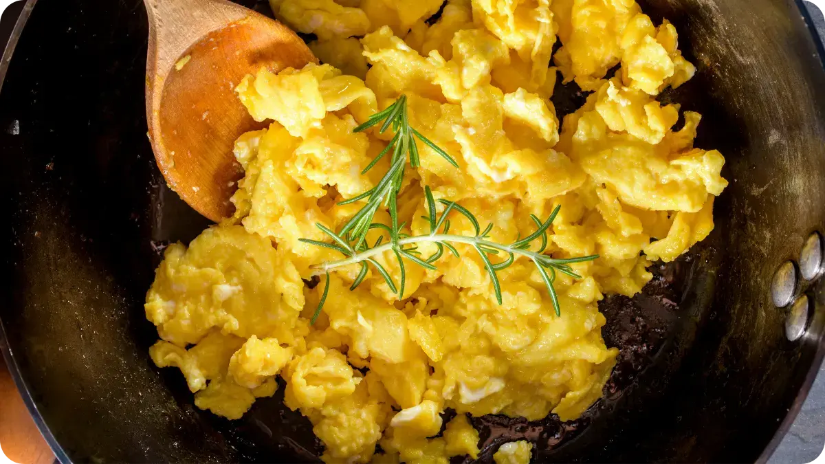 Scrambled egg with rosemary on top, being stirred with a wooden spoon.